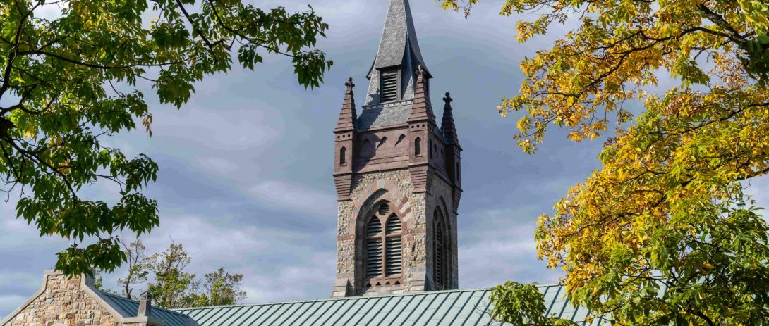 The main subject of the image is a tall church steeple with a pointed spire and decorative architectural details. The action is static, as the steeple stands still without any movement. The setting is outdoors during the day, surrounded by green and yellow foliage, indicating it might be early autumn, under a partly cloudy sky.
